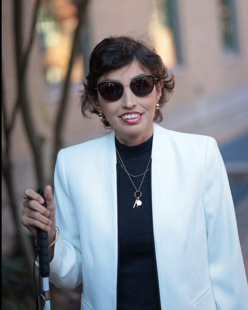 Ana standing outdoors holding a white cane, wearing sunglasses and a white blazer over a dark top, smiling confidently with a blurred background behind her.