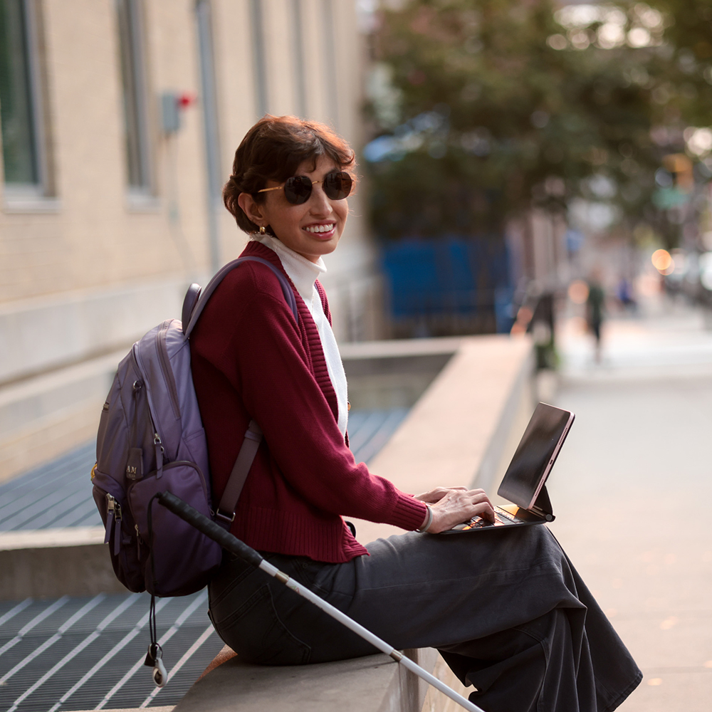 Ana sitting on an urban sidewalk with a white cane, using a laptop on her lap and smiling, with buildings and people blurred in the background